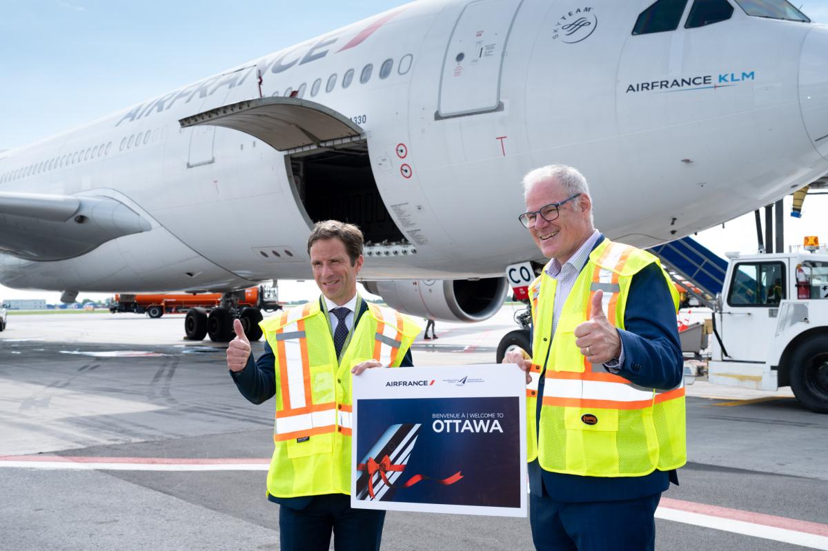 Deux hommes portant des vestes de haute visibilité lèvent le pouce en tenant une pancarte portant les logos d'Air France et de l'Administration de l'aéroport et indiquant Bienvenue à Ottawa / Welcome to Ottawa, avec un avion en arrière-plan.