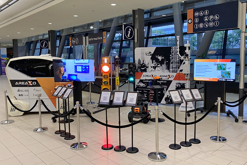 A shuttle vehicle, a robot, a drone, a traffic light, two monitors, and other pieces of technology on tiled floor behind a stanchion. A series of QR codes mounted on waist-height stands in the foreground. Promotional branded banners in the background.