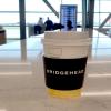 Close up of disposable coffee cup with a protective sleeve printed with the Bridgehead logo, on a counter. 